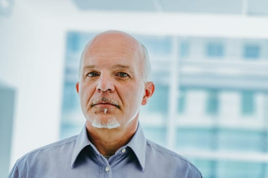 In front of a large window, an elderly man with a bald head and grey goatee stands, wearing a blue shirt. His expression is serious, reflecting a contemplative state