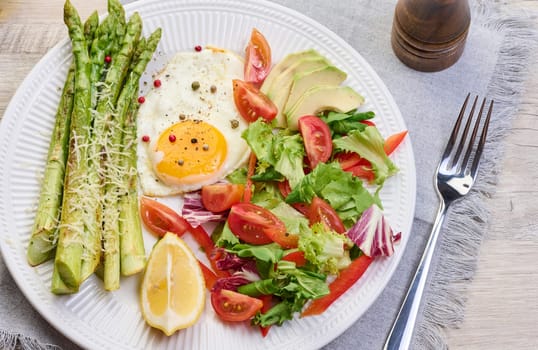 Round plate with cooked asparagus, fried egg, avocado and fresh vegetable salad on the table, top view
