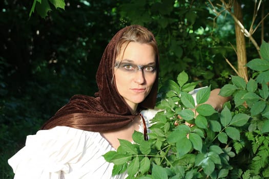 A woman as a medieval huntress in a dress with a hood hides in the forest