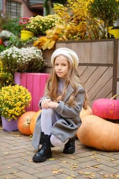 Little girl in a beret and wool coat sits on squats next to colorful pumpkins