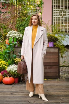 Elegant woman with long hair wearing beige wool coat, holding brown handbag and posing on autumn day