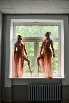 Two graceful ballerinas in transparent long dresses stand on the windowsill in the corridor