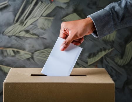 Ballot box with hand voting on a blank ballot paper against a light background.