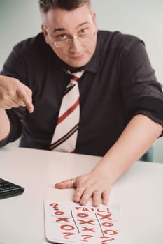 Seated man in glasses, dark shirt, and striped tie points at a tic-tac-toe sheet marked 'FAIL,' showing disappointment, indicating failure or frustration in an office environment