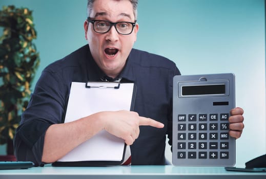 Man in dark shirt and glasses, holding a clipboard with one hand and pointing at a large calculator with the other, expresses surprise, seated at a desk with a plant and office equipment