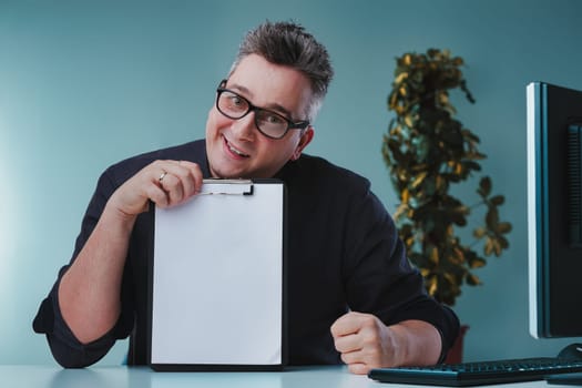 Man in dark shirt and glasses, holding a clipboard with a blank sheet, shows an uncertain and skeptical expression, seated at a desk with a plant and computer, conveying hesitation