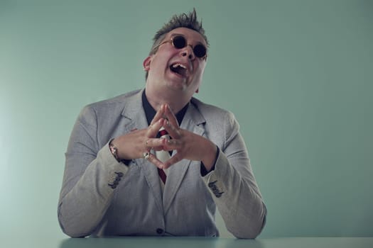 Mad evil Joyful executive in grey blazer and sunglasses laughs uproariously, showing a spirited personality at his desk