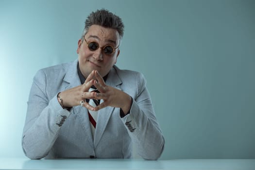 Businessman in grey suit smirks confidently behind desk, fingers interlocked, wearing round sunglasses and displaying various rings