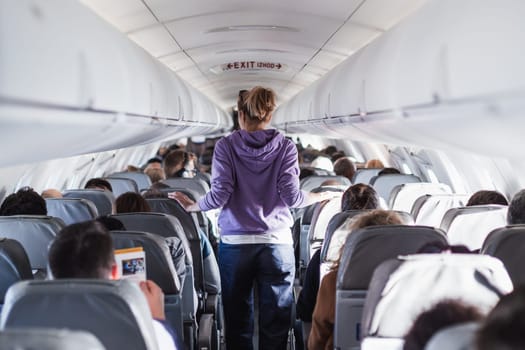 Interior of airplane with passengers on seats and female traveler walking the aisle. Commercial economy flight service concept