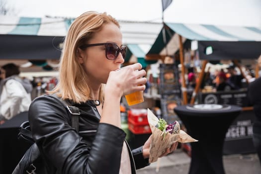 Beautiful young woman holding delicious organic salmon vegetarian burger and drinking homebrewed IPA beer on open air beer an burger urban street food festival in Ljubljana, Slovenia