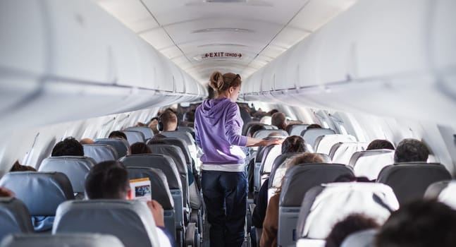Interior of airplane with passengers on seats and female traveler walking the aisle. Commercial economy flight service concept