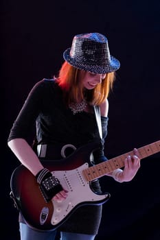 Smiling young woman with auburn hair plays an electric guitar on stage. She wears a sparkling hat, dark glittery top, jeans, and fingerless gloves, exuding confidence and joy in her performance