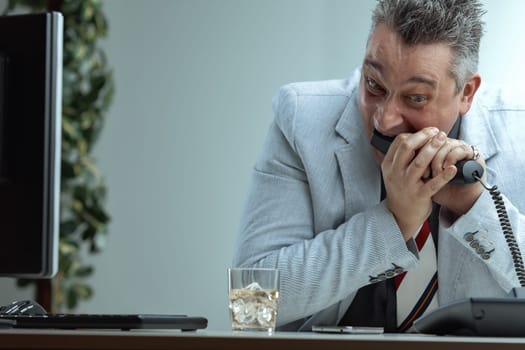 Wearing a light suit and striped tie, the man eats a phone with a frustrated expression. A glass of whiskey on his desk indicates the stress of managing public relations difficulties