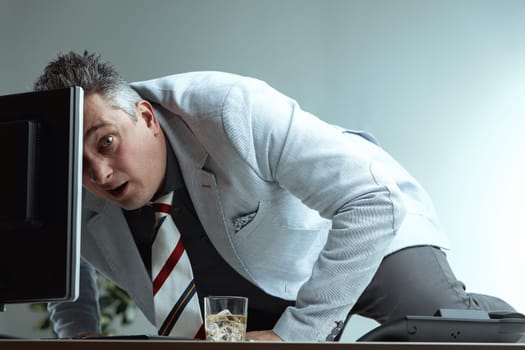 man, dressed in a light suit and striped tie, appears startled while leaning over his desk. His surprised expression and awkward posture, along with a whiskey glass, imply he was caught doing somethin