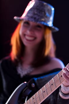 Young woman with auburn hair, in a sparkling hat and dark glittery top, plays an electric guitar. Her smile and confident pose on stage convey her enthusiasm for performing