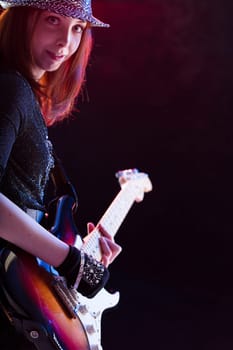 Smiling auburn-haired woman wearing a glittery hat and dark top plays an electric guitar. Her confident stance and joyful expression on stage emphasize her love for music