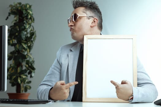 Confident man with spiky gray hair and sunglasses points at a blank whiteboard. His light gray suit and black shirt, combined with his smug look, suggest hes offering something exceptional