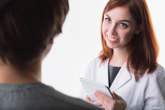woman with auburn hair, wearing a white lab coat, holds a tablet and gazes gently at a patient. Her attentive expression highlights her professional competence in healthcare