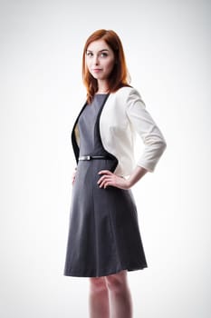 young woman with auburn hair, dressed in a white blazer and dark dress, stands confidently with hands on hips. Her poised demeanor and professional attire convey authority and competence