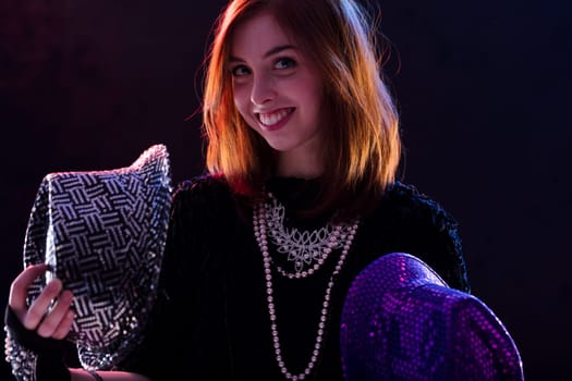 Smiling auburn-haired woman holds a sequined black hat and a purple hat in a dimly lit environment. Her layered necklaces and confident expression highlight her playful, rockstar personality