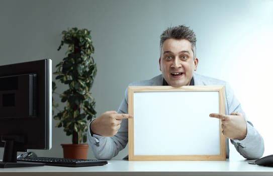 man in a light suit and striped tie, smiling widely, points at a blank whiteboard. His enthusiastic expression suggests he is eager to present or communicate an important message