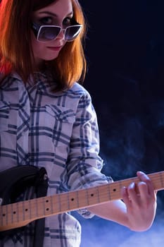 young woman with auburn hair, wearing a plaid shirt and sunglasses, plays an electric guitar with a bright smile. Her energetic pose highlights her enthusiasm and confidence in performing