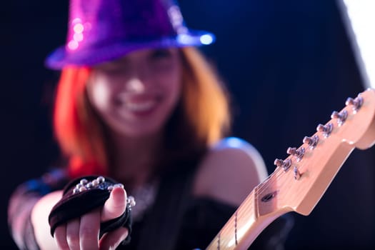 Young woman with auburn hair, sparkling hat, and dark glittery top plays an electric guitar with a smile. Her confident and joyful performance on stage showcases her musical talent
