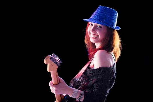 woman with auburn hair and a glittery hat plays an electric guitar with a smile. Dressed in a dark top and jeans, she exudes confidence and joy on stage