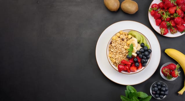 Granola with strawberries, kiwi, banana and blueberries in a round plate on a black table. Healthy and tasty food, top view. Copy space