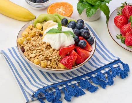 Granola with strawberries, kiwi, banana and blueberries in a round plate on a white table. Healthy and tasty food, top view