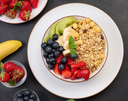 Granola with strawberries, kiwi, banana and blueberries in a round plate on a black table. Healthy and tasty food, top view