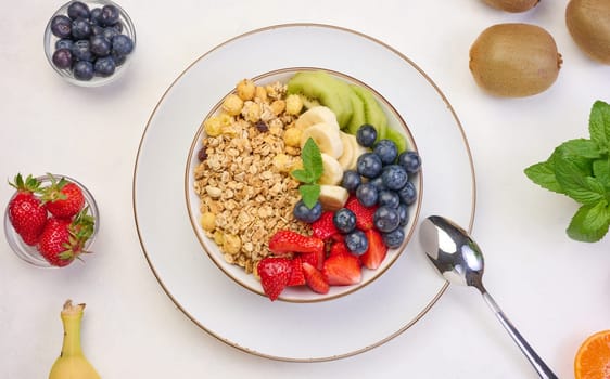 Granola with strawberries, kiwi, banana and blueberries in a round plate on a white table. Healthy and tasty food, top view