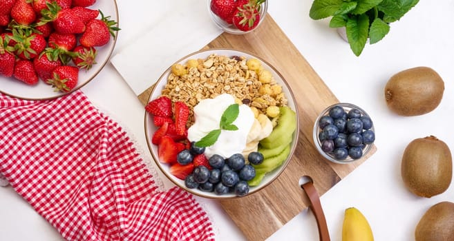 Granola with strawberries, kiwi, banana and blueberries in a round plate on a white table. Healthy and tasty food, top view