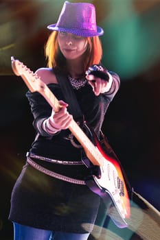 joyful young woman with red hair, in a glittering black ensemble and sequined hat, skillfully plays her electric guitar, stage lights casting colorful reflections around her