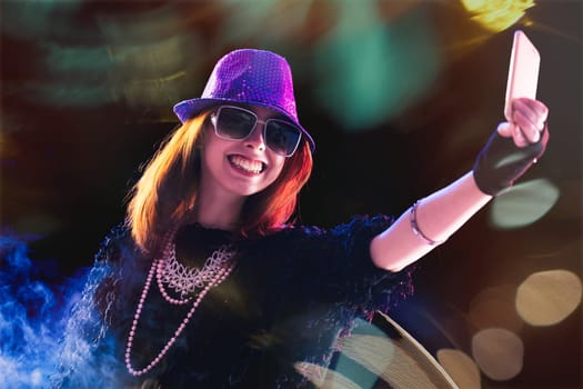 Smiling brightly, a red-haired woman in a sequined hat and sunglasses uses her phone, immersed in a virtual event, her stylish outfit glowing under the vibrant stage lights