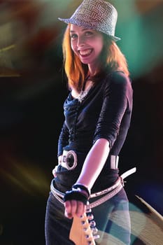With a radiant smile, a red-haired young woman in a sparkling black outfit and sequined hat plays an electric guitar, her joy evident as colorful stage lights illuminate her