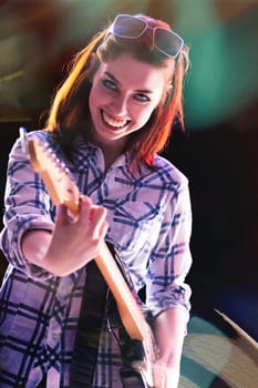 Under vivid stage lights, a young woman with red hair and a plaid shirt passionately plays her electric guitar, her wide smile and sunglasses on her head showcasing her joy