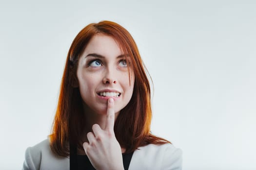 Thoughtful redhead with an inquisitive expression, gently touching her chin, dressed in a sophisticated white blazer