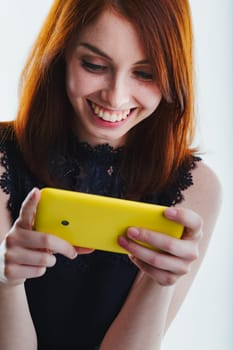 young red-haired woman, dressed in a black sleeveless top, engages with her yellow smartphone, smiling widely as she interacts with the device in a simple, plain setting