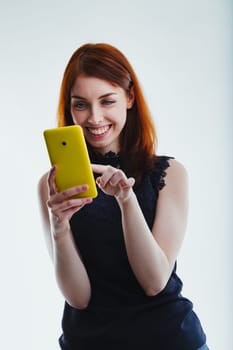 Enthusiastically using her yellow smartphone, a red-haired woman in a black top smiles broadly, her fingers moving across the screen against a clean, uncluttered background
