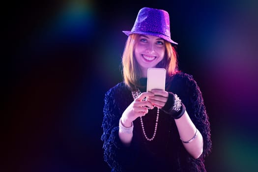 red-haired young woman in a purple sequined hat and sunglasses uses her phone, smiling confidently. Wearing a stylish black top and jeans, she is surrounded by stage lights and fog