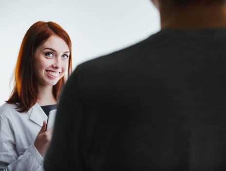 ed-haired female doctor in a white lab coat smiles warmly while holding a tablet, engaging with a patient. Her professional yet friendly demeanor underscores the importance of compassionate healthcare