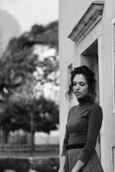 young woman stands near a building, looking coyly down. She wears a brown turtleneck and a grey skirt. The black and white photo features a blurred background with trees and buildings