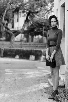 young woman stands near a building, appearing to wait for someone. She wears a brown turtleneck and grey skirt, holding a clutch. The black and white photo features a blurred background of trees