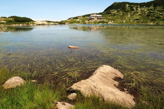 View over the Fish Lake, Ribnoto Ezero, water plants, grass can be seen below the smooth surface, the Seven Lakes Chalet can be seen on the shore in the background reflecting in the water, Rila National Park, close to Sofia, Bulgaria 2022