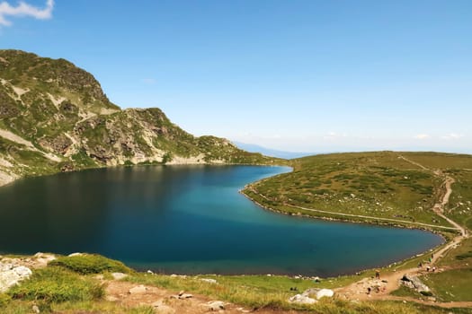 Spectacular view onto the deep blue Kidney, Babreka Lake, one of the Seven Rila Lakes, situated on a plateau in the Rila National Park, a few people are on the hiking trail right next to it, close to Sofia, Bulgaria 2022