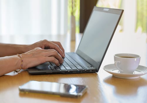 Female working on laptop at home. Woman fingers typing on keyboard while sitting at desk with mobile phone and a cup of coffee next to laptop