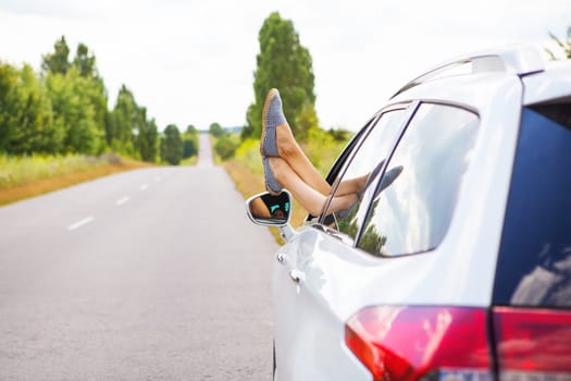 A young girl driving a car, pulling her legs out of the window, resting behind the wheel. Road trip