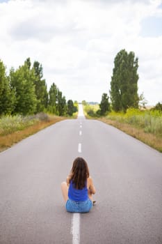 Attractive girl sitting on the asphalt and posing in the middle of the asphalt road near the field, copy space for content.