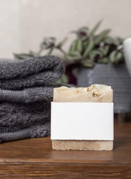 Soap bar with blank label near grey folded towels, green plant and vessel basin on brown wooden countertop in beige bathroom, close up, brand packaging mockup. Natural handmade hygiene product
